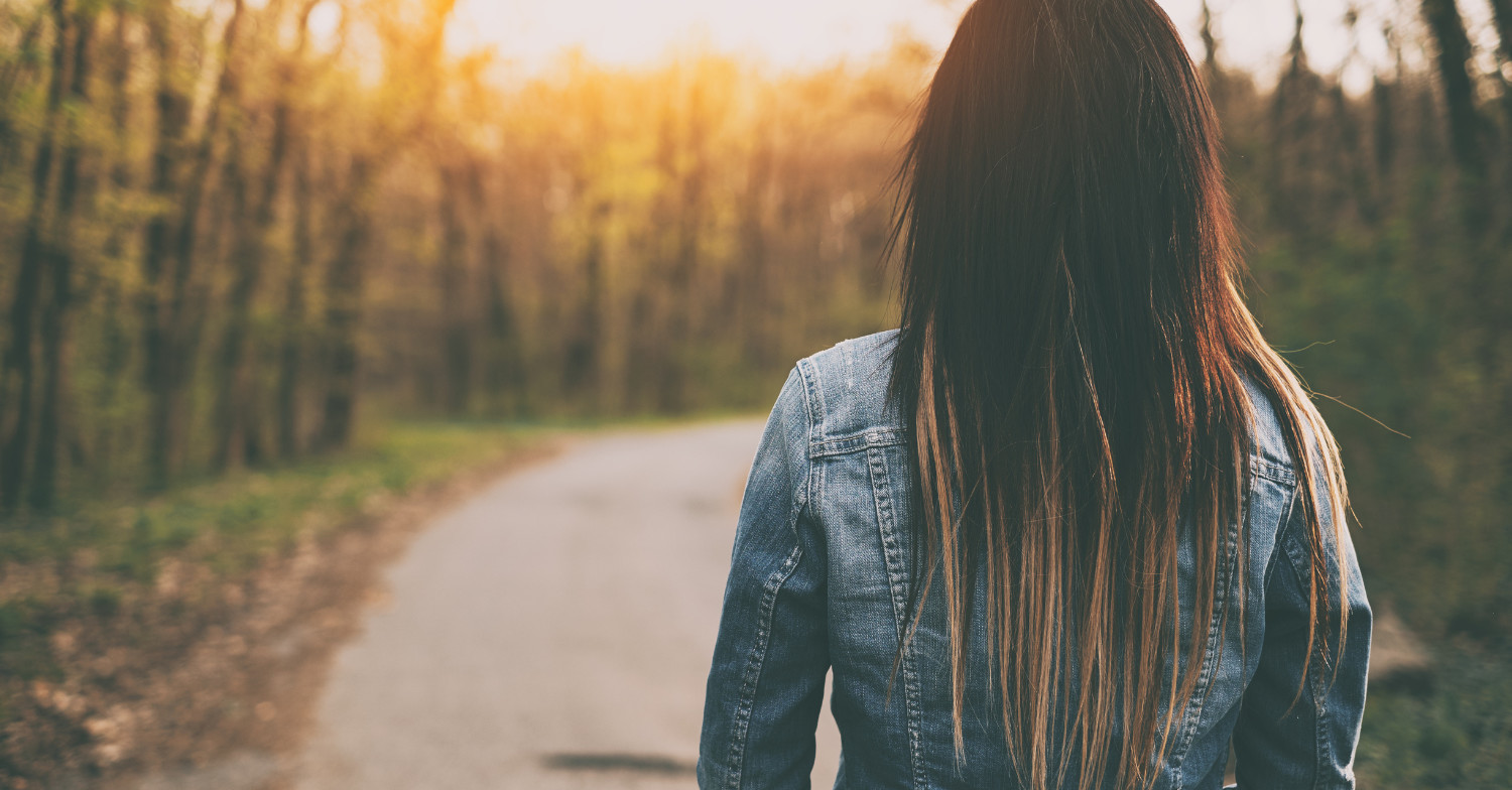 A brunette female wearing a jean jacket, walking away from view along a paved tree-lined pathway while the sun is setting. Alone, but moving forward peacfully vibes.